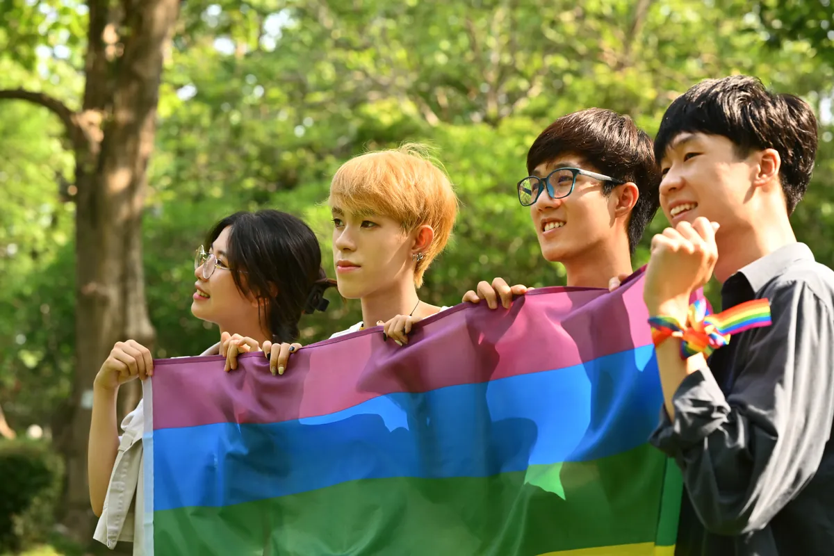 Four friends in a sunny, tree-lined park smile as they hold up a large rainbow flag together, one wearing a rainbow ribbon on his wrist